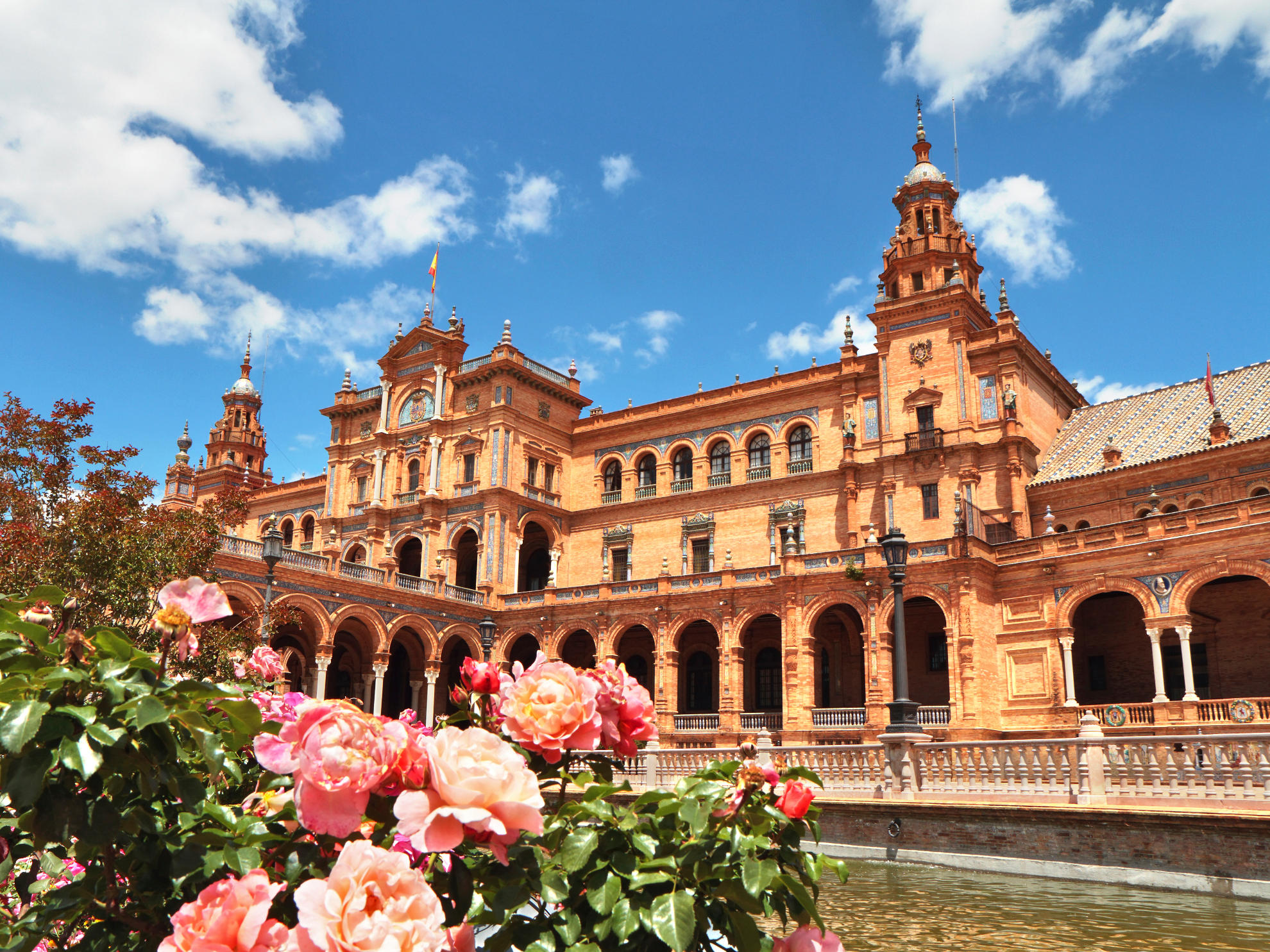 A picturesque view of the Plaza de España in Seville, Spain. The ornate building, with its orange facade, stands majestically against a clear blue sky. A tranquil waterway and colorful flowers in the foreground complete the idyllic scene.